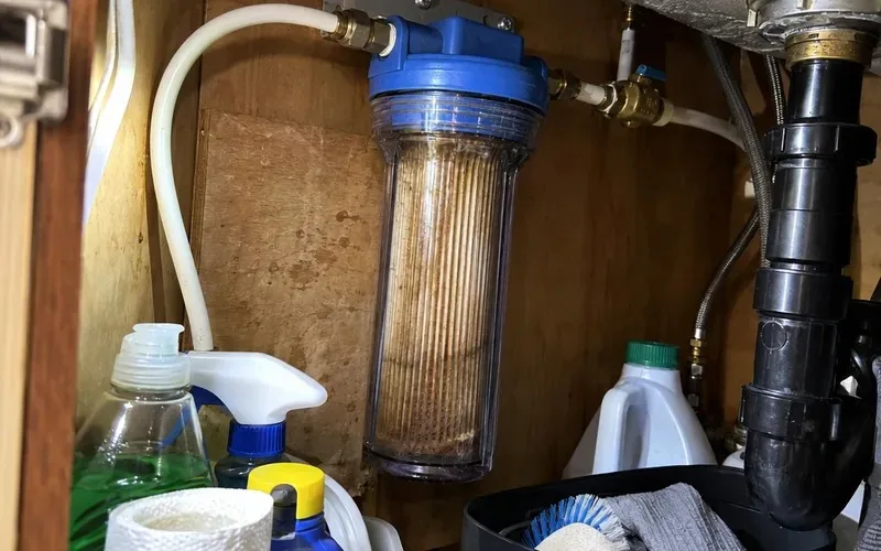 Clear 10 inch filter housing under a sink showing a brown, sediment-clogged cartridge
